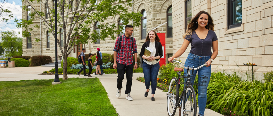Woman on bicycle and two other students walking behind her on sidewalk in front of campus