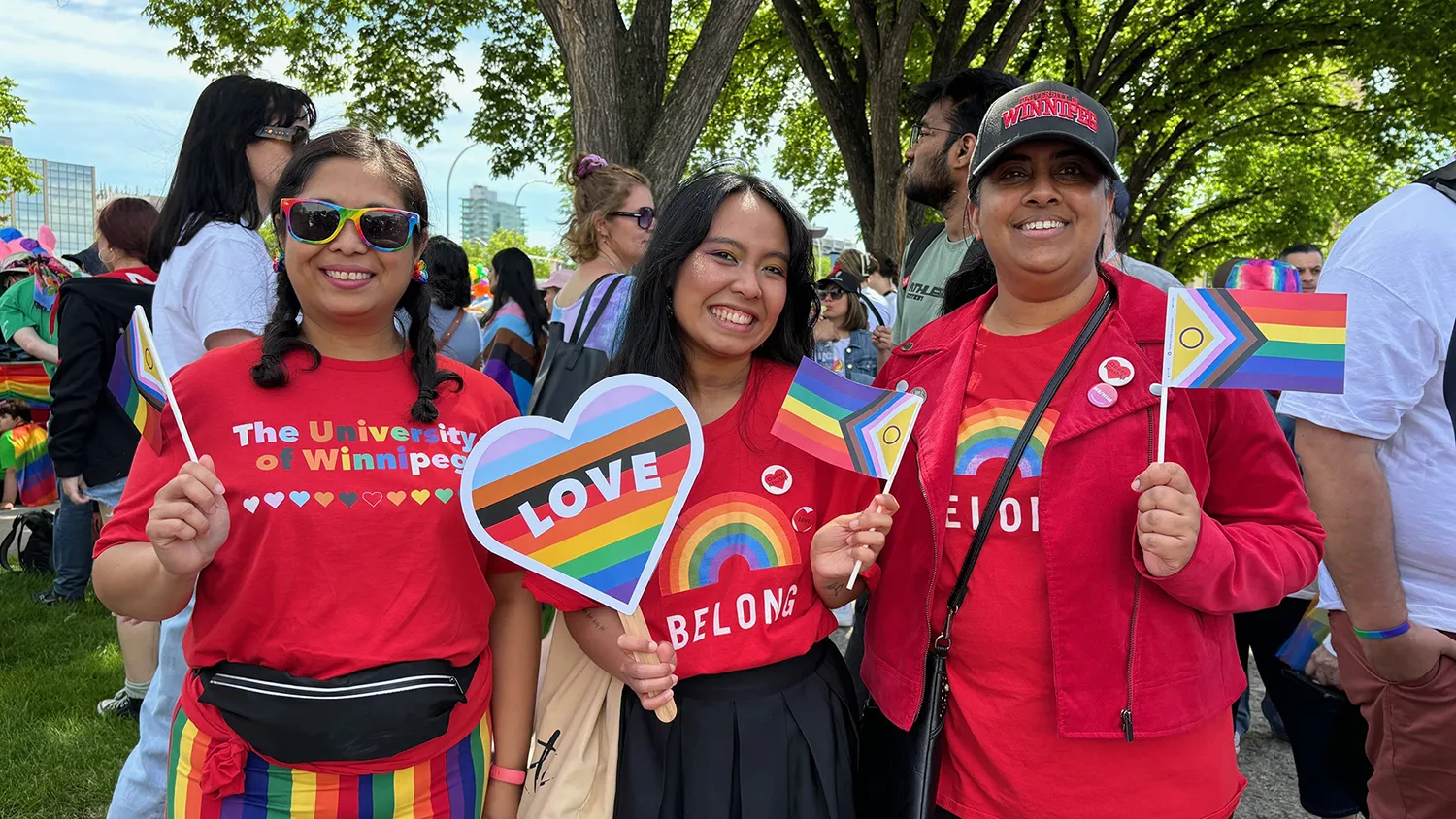 Three people holding rainbow flags and dressed in pride t-shirts smile at the camera.
