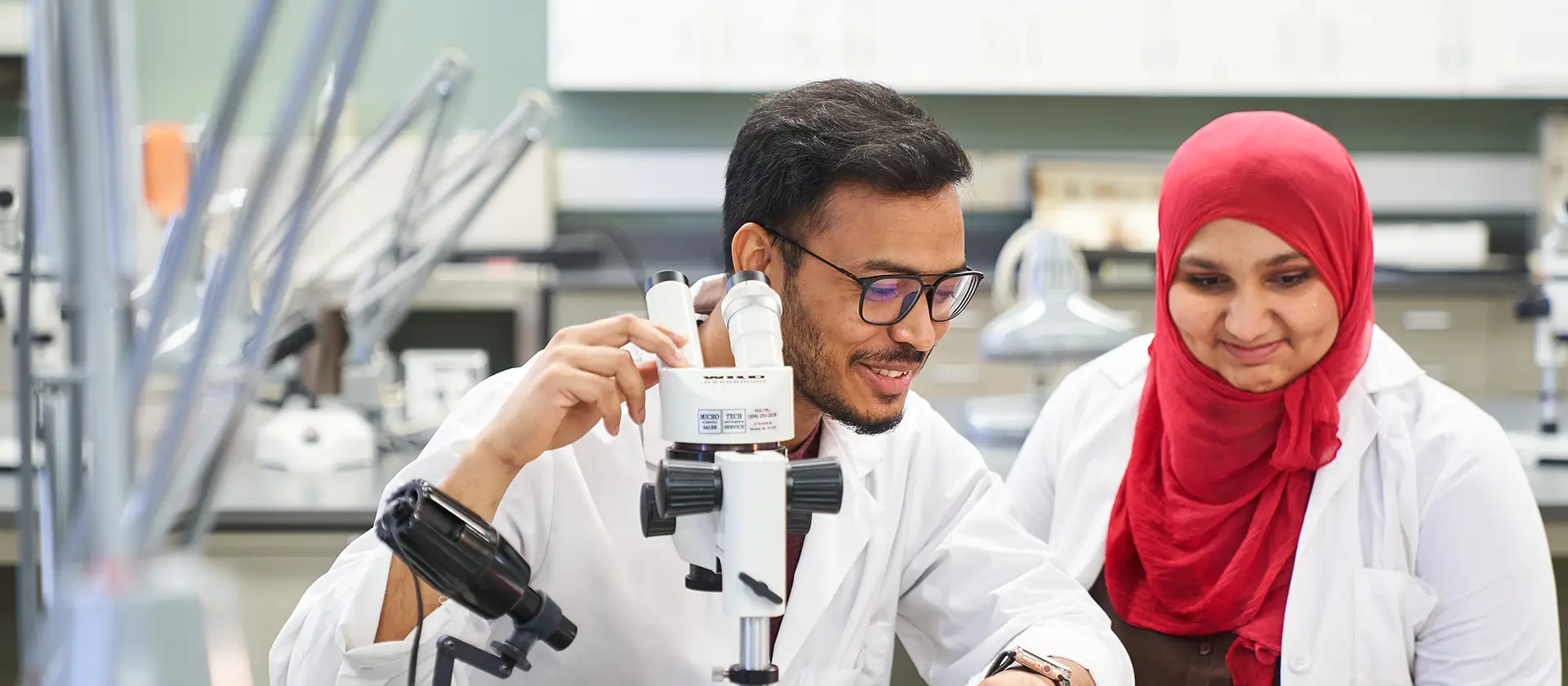 Two students in a lab, one at a microscope.