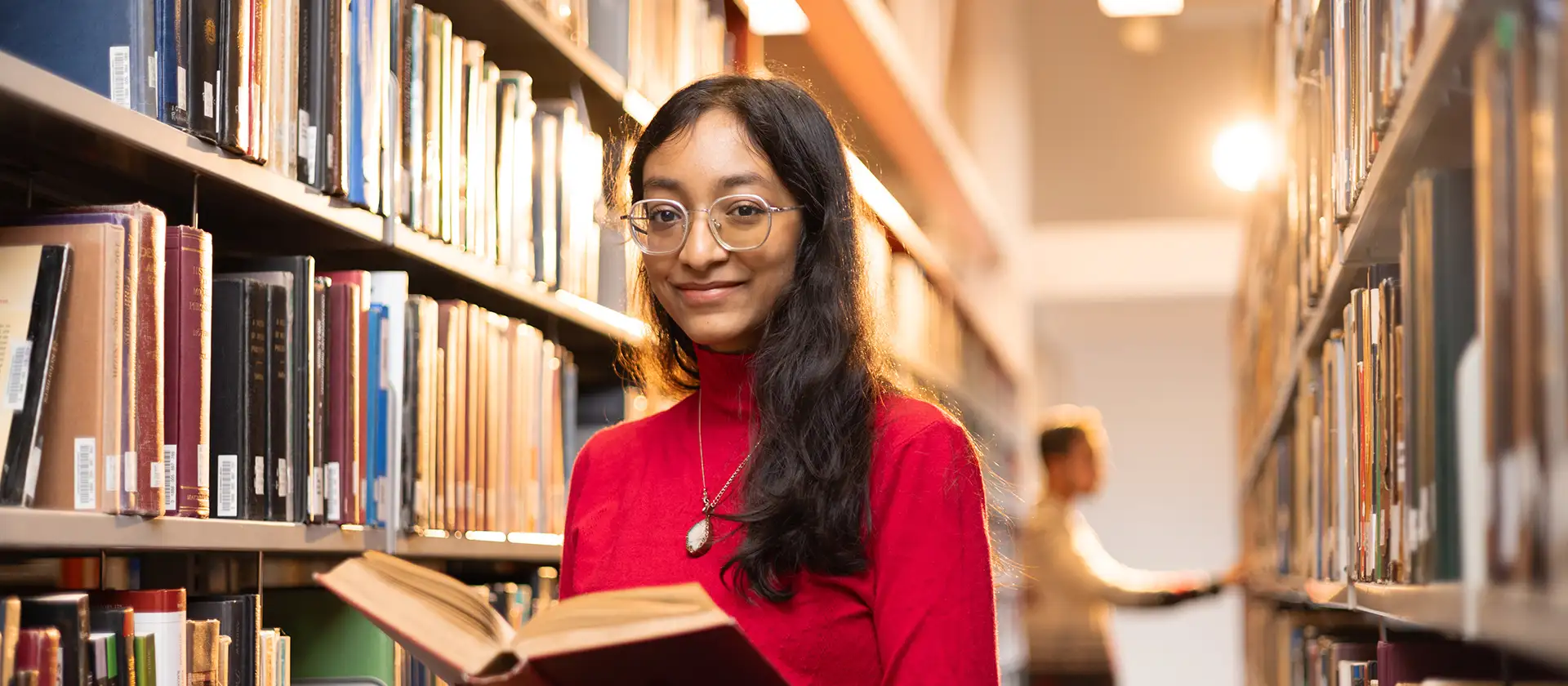 Student in the library in red sweater and gold glasses. She is holding a book and smiling at the camera.