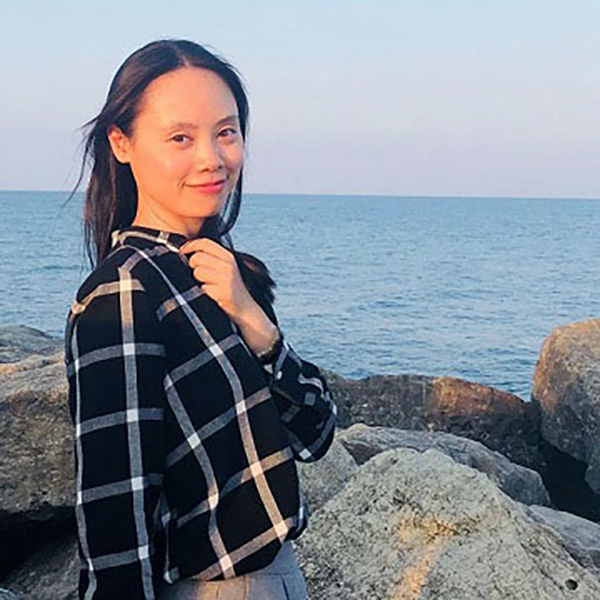 Portrait of a young woman with long dark hair, wearing a plaid shirt, standing in front of rocks and water.