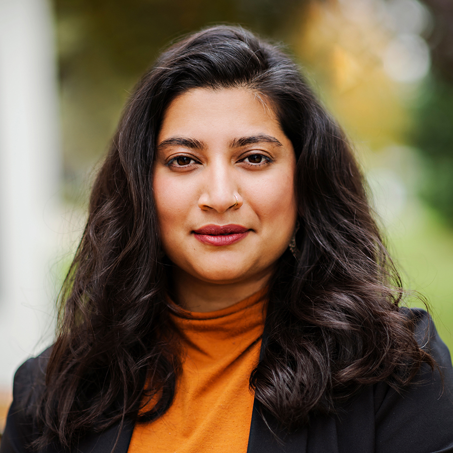 Close-up portrait of a young woman with long dark hair and a nose ring, wearing an orange turtleneck and black blazer.