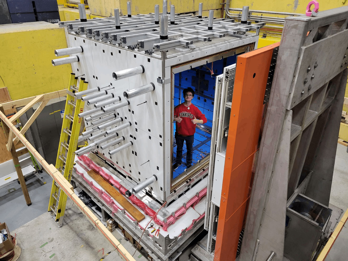 Person in a red shirt and dark pants standing in a large metal box inside a large room. The box has metal pipes coming out of the sides and top.