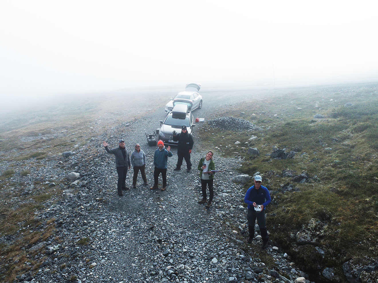 Taken from above, this image shows six people staring up at the camera and waving. Two cars with open trunks sit behind the people, who are standing on a rocky paved road with fog behind them.