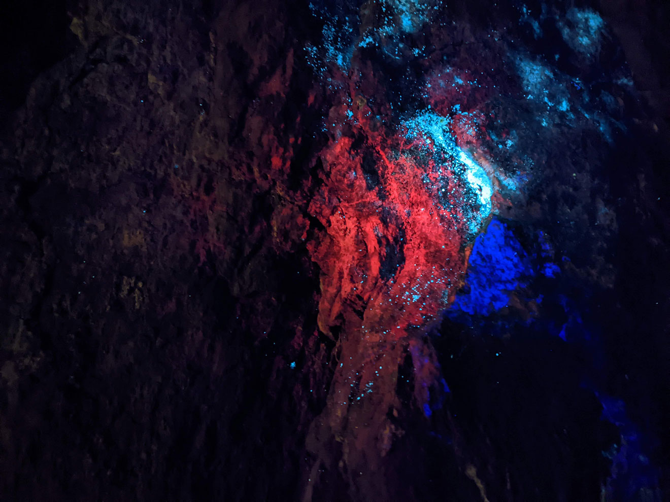 An uneven rock wall glowing with patches of bright red, blue and turquoise.