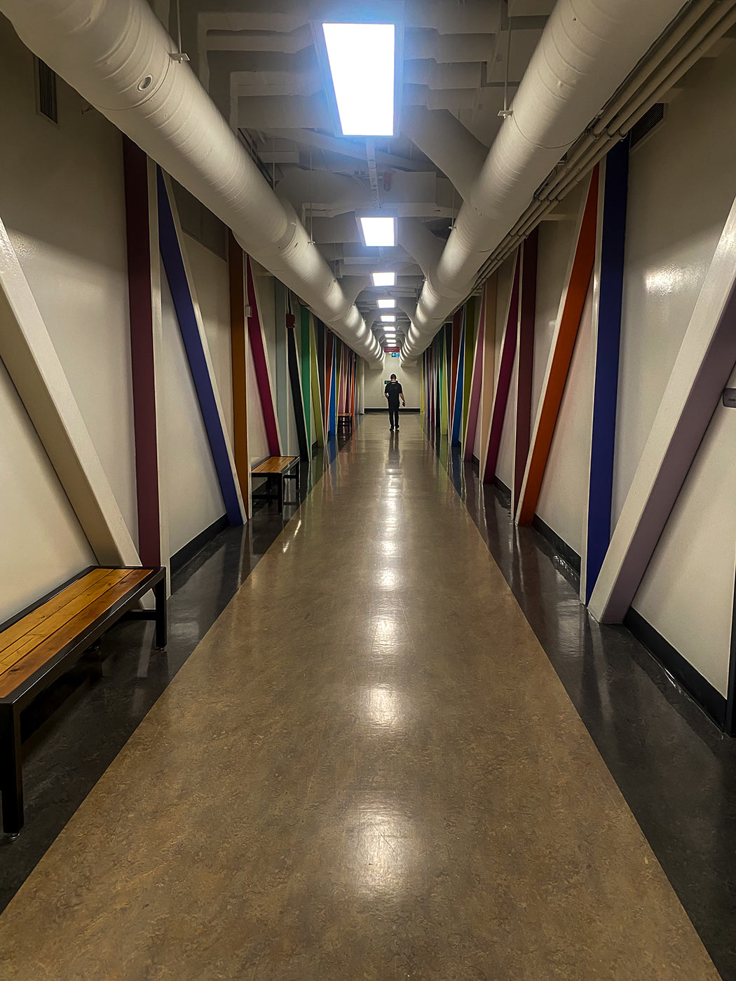 A long corridor with polished floors, overhead fluorescent lights, and exposed white ceiling pipes. The walls are lined with colourful diagonal beams.