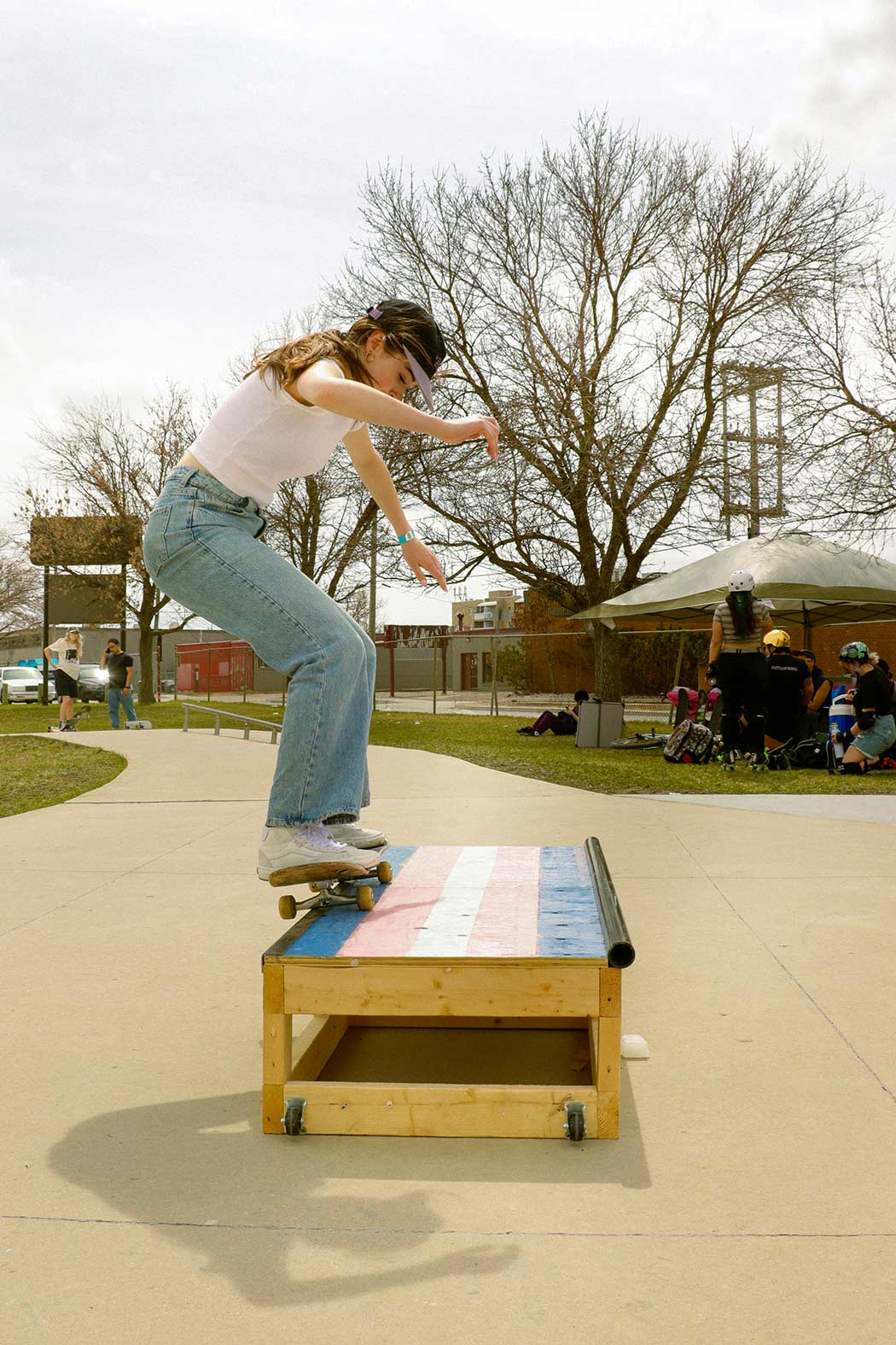 A woman in an outdoor skatepark, wearing a white shirt and jeans balances on her skateboard on top of an obstacle decorated with the trans pride flag.