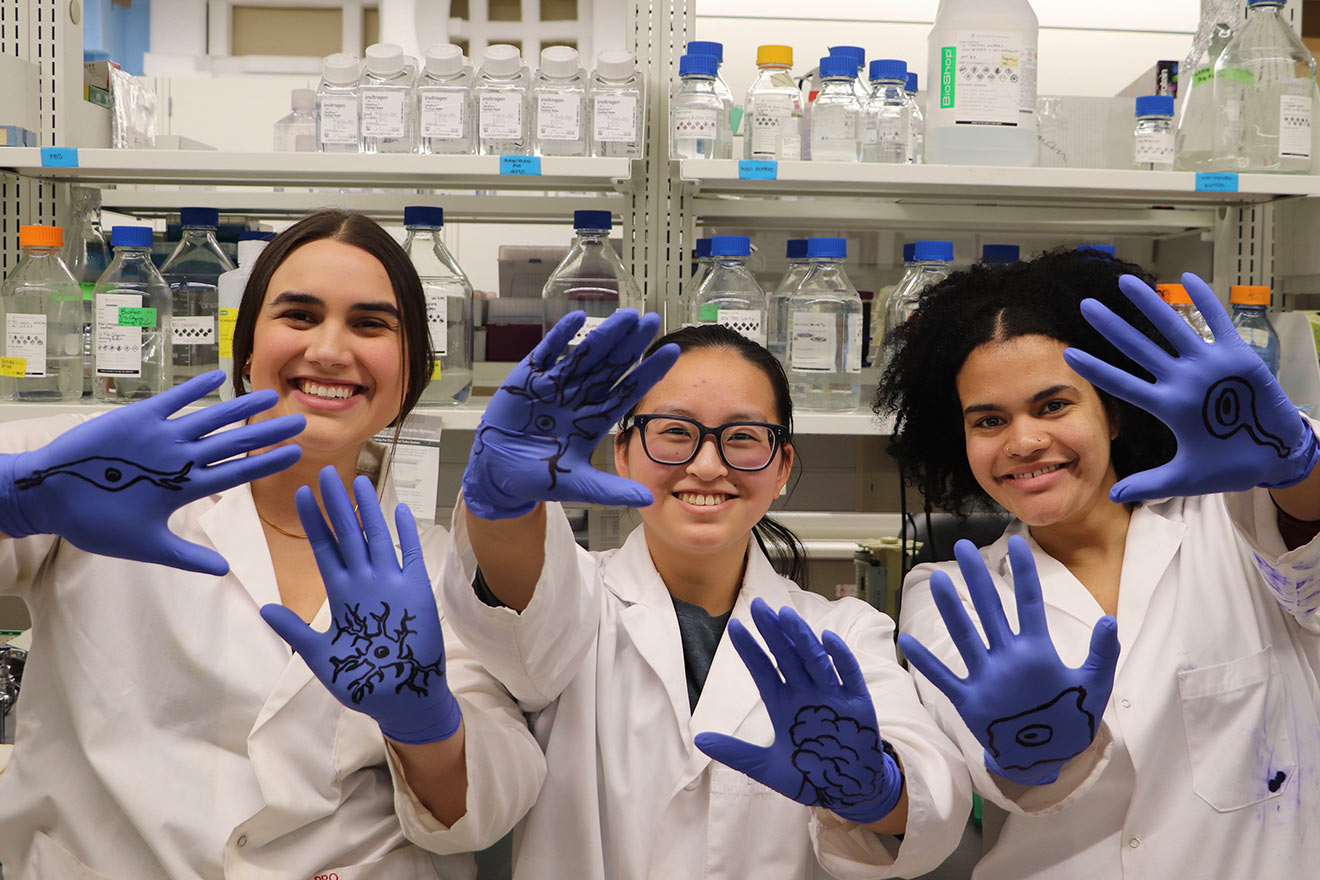 Three female scientists smiling at the camera, wearing blue gloves with pictures drawn on them in sharpie. Shelves with reagent bottles in background.