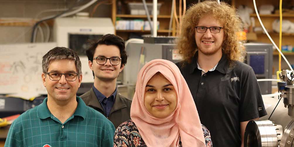The photo is a group shot of Dr. Russell Mammei, Thomas Hepworth, Abeer Zahra, and Rylen De Vries standing in a research lab staring directly at the camera