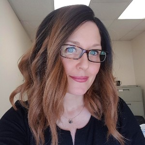 Stacey is smiling, she has mid length wavy brown hair. There is a white wall behind her.