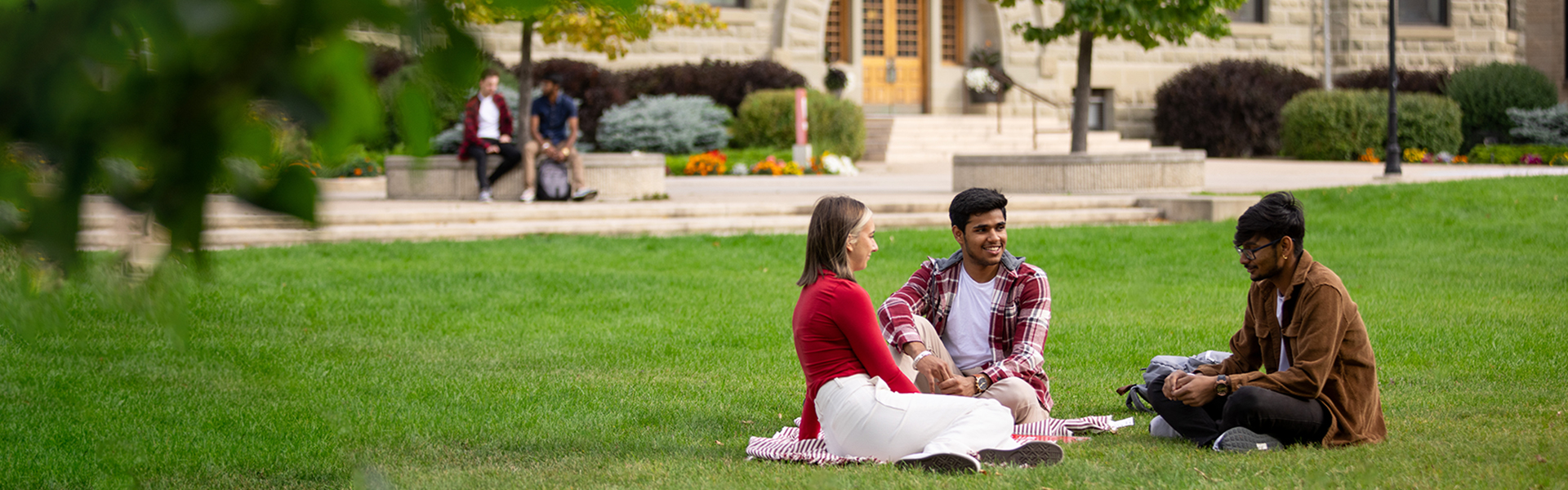 Three students sit on the lawn in front of Wesley Hall on UWinnipeg's campus.
